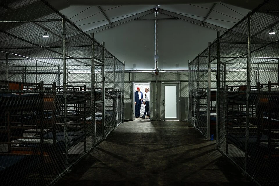 Rows of bunk beds line the interior of the so-called “Alligator Alcatraz” detention facility in the Florida Everglades, a repurposed training center now designated for holding immigrants. President Donald Trump appears in the background during a July 1, 2025, visit to the site.?w=200&h=150